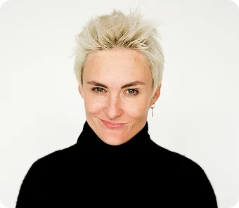 Portret kobiety z krótkimi blond włosami, ubranej w czarny golf, lekko się uśmiechającej na jasnym tle.